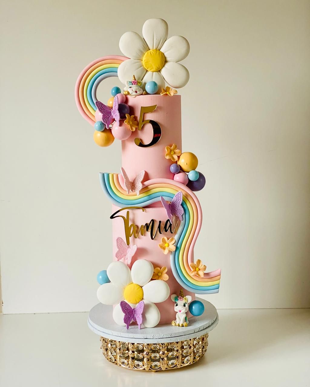 A two-tier pink celebration cake with a pastel rainbow, giant sugar daisies and butterflies